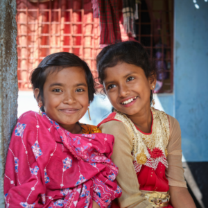 Kaksi pientä tummahiuksista tyttöä istuu vierekkäin värikkäissä vaatteissa ja hymyilee.